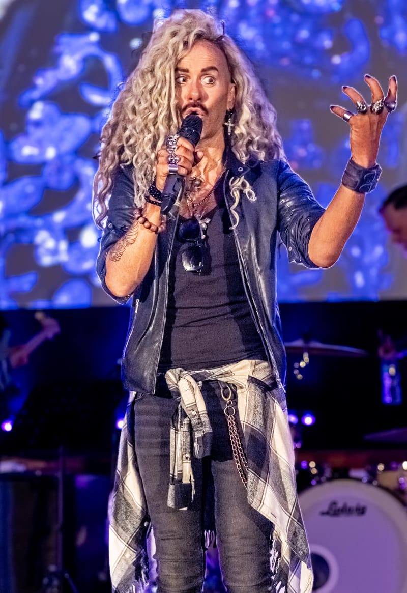 Performer with long blonde wavy hair singing into microphone on stage with blue lighting backdrop.