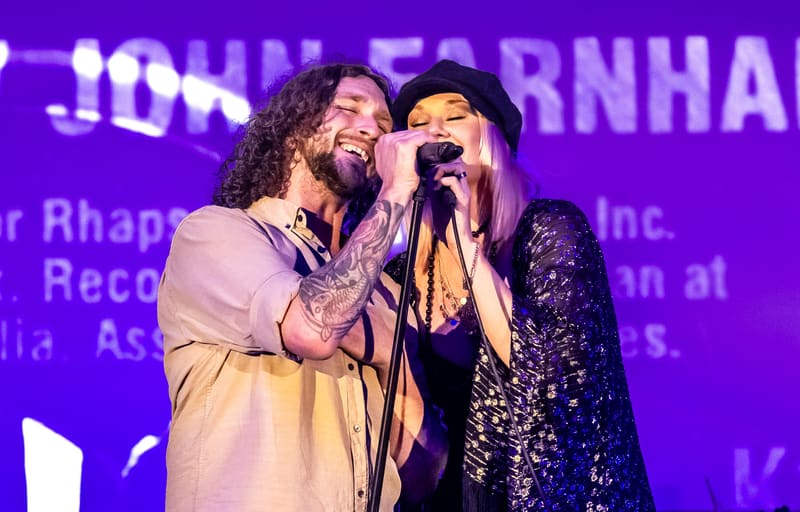 Two performers share a microphone on stage beneath purple lighting during a collaborative musical performance.