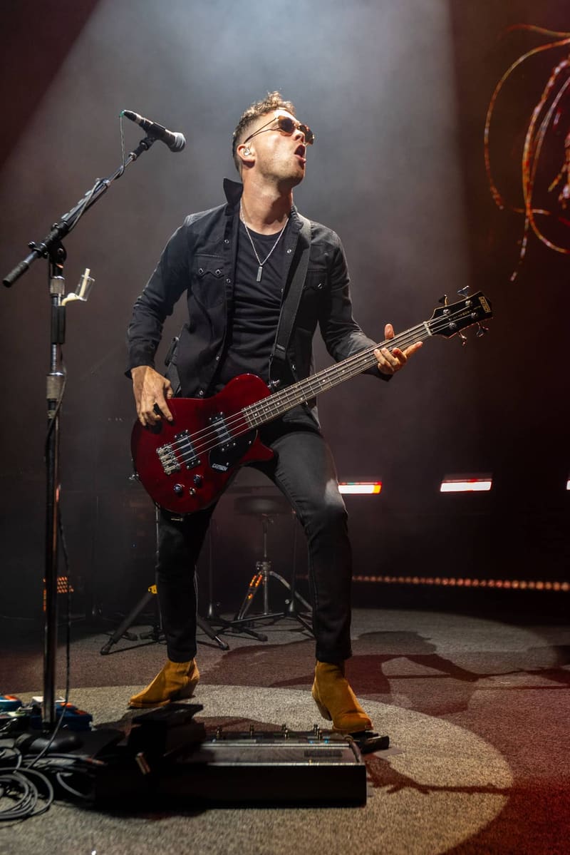 Bassist playing a red electric bass guitar on stage under dramatic spotlighting during a live rock performance.