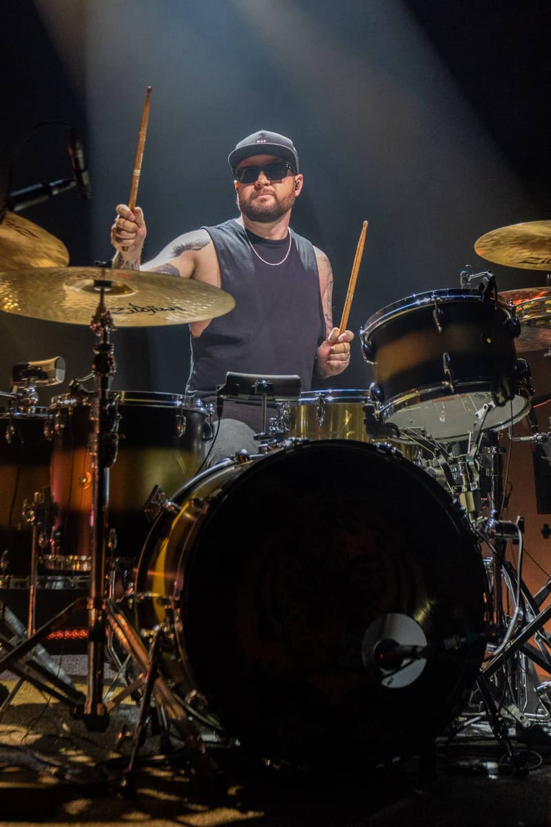 Drummer in sunglasses and cap performing at drum kit under warm stage lighting during Royal Blood concert.