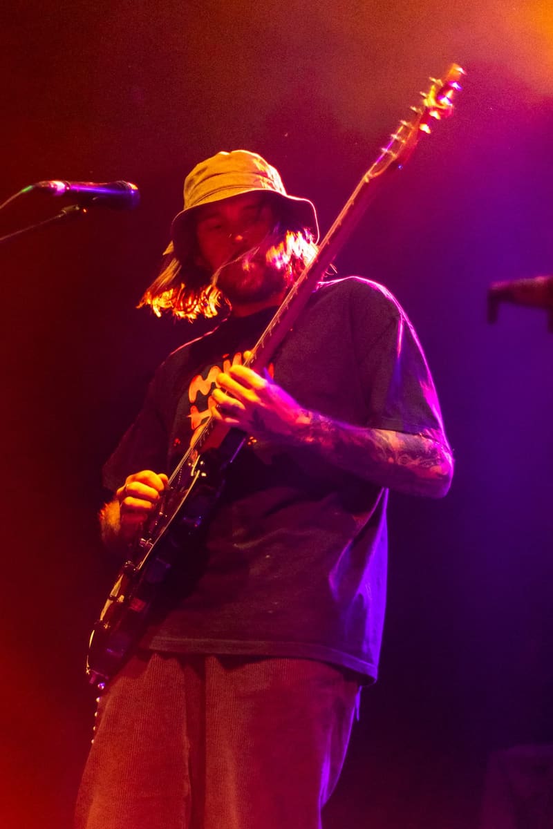 Guitarist wearing a hat performs on stage bathed in warm orange and purple stage lighting.