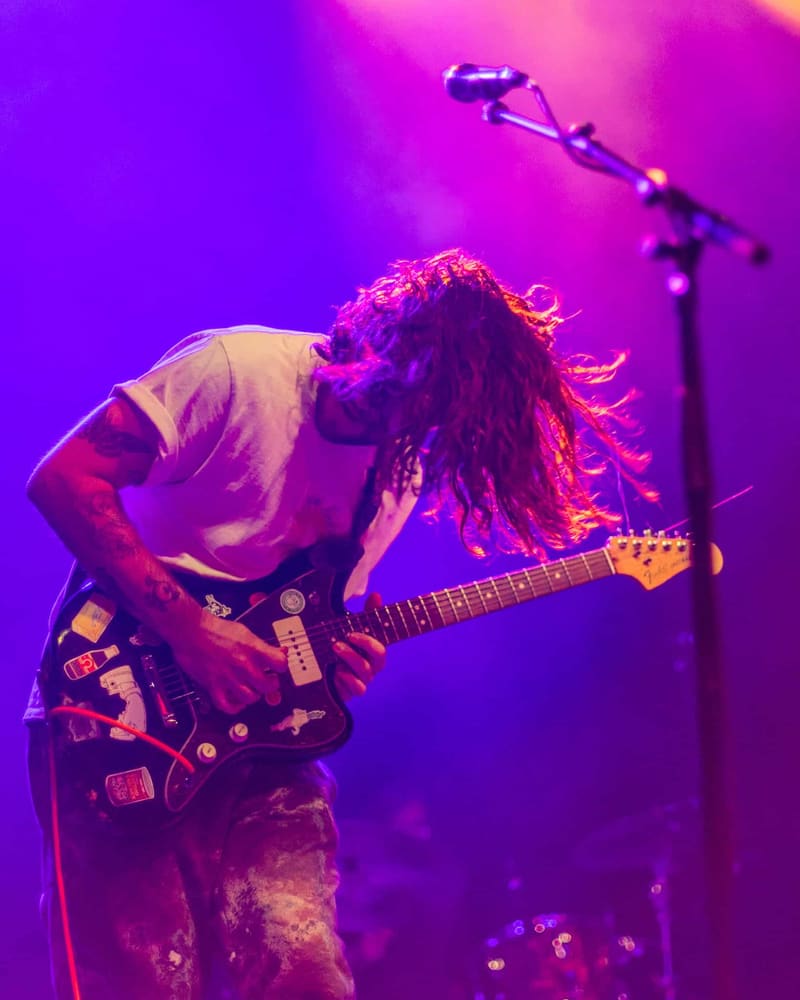 Guitarist with long hair bent over instrument, performing under vibrant purple and pink stage lighting.