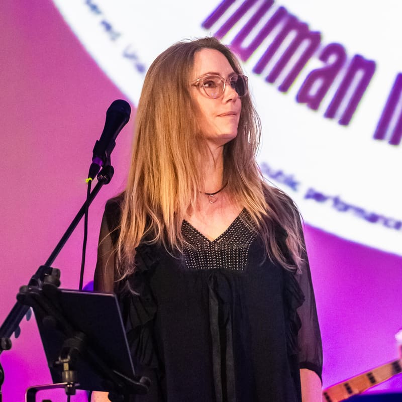 Woman with long hair and glasses speaking at a microphone against a vibrant purple and white backdrop.