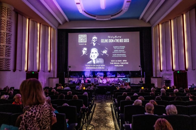 Theatre audience seated facing a large screen displaying performance imagery, with live musicians on stage beneath purple lighting.