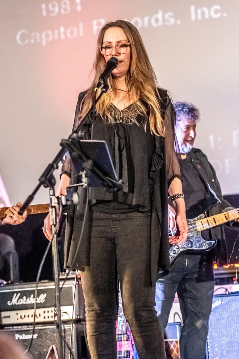 Singer with long blonde hair and glasses performs at microphone whilst band members play instruments behind her on stage.