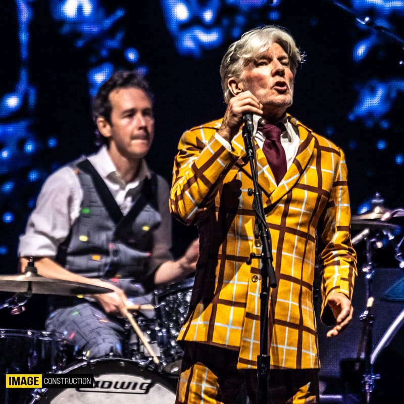 Singer in bright yellow plaid jacket performs at microphone with keyboardist during Split Enz concert.