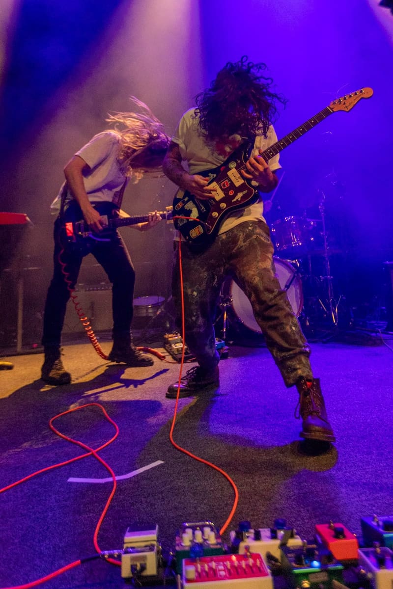 Two musicians perform on stage bathed in purple light, one playing guitar while the other sings, with effects pedals visible in the foreground.