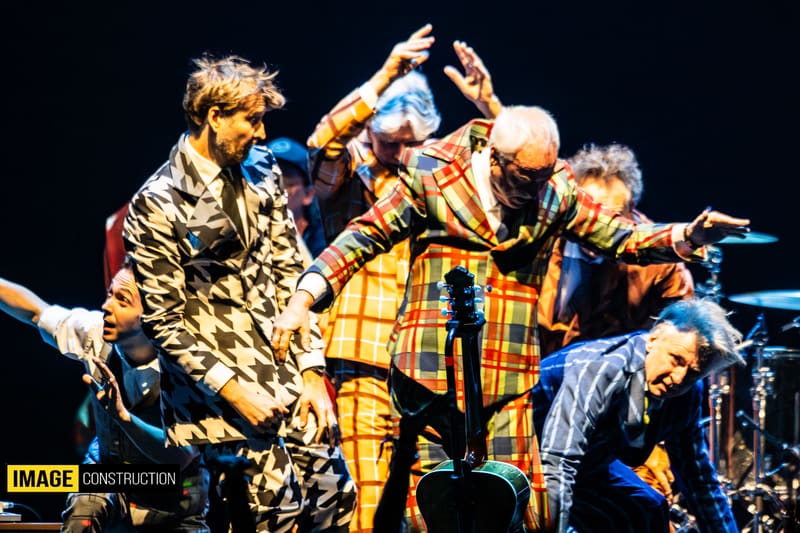 Split Enz band members performing on stage in colourful patterned clothing under warm stage lighting.