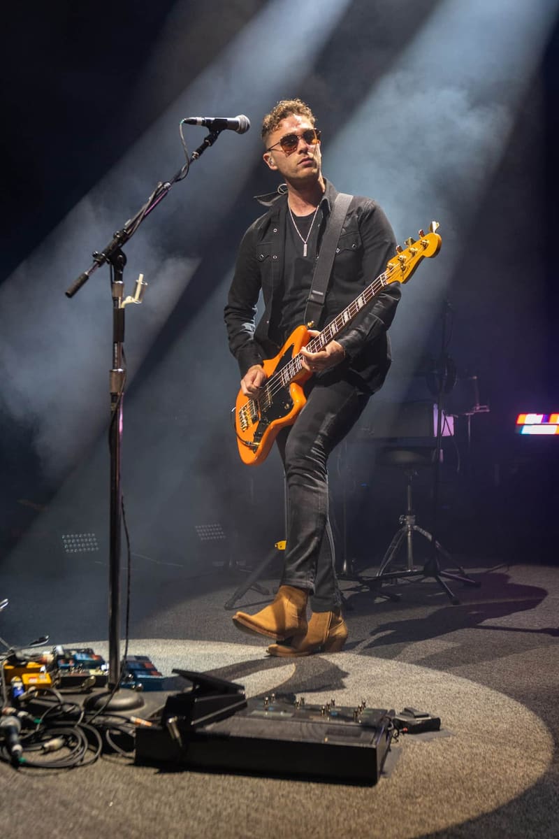 Guitarist in black jacket and sunglasses performs with orange electric guitar under dramatic stage lighting.