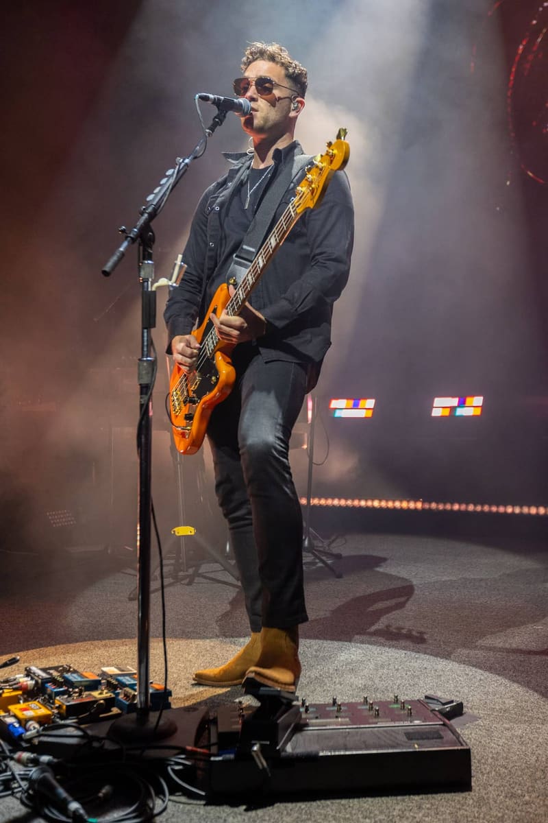 Musician performing with orange electric guitar at a microphone on a dimly lit stage with atmospheric lighting.