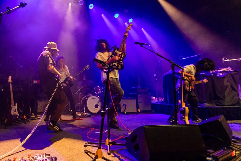 Four band members perform on stage bathed in purple and blue stage lighting.