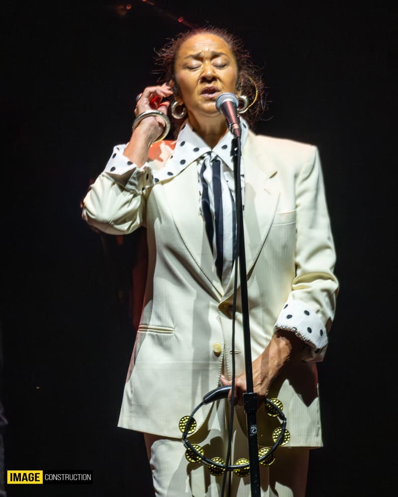 Singer in cream blazer and polka-dot collar performing at microphone on stage.
