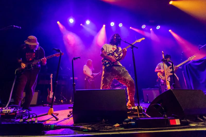 Psychedelic Porn Crumpets perform on stage bathed in purple and pink stage lighting, with band members playing guitar and other instruments.