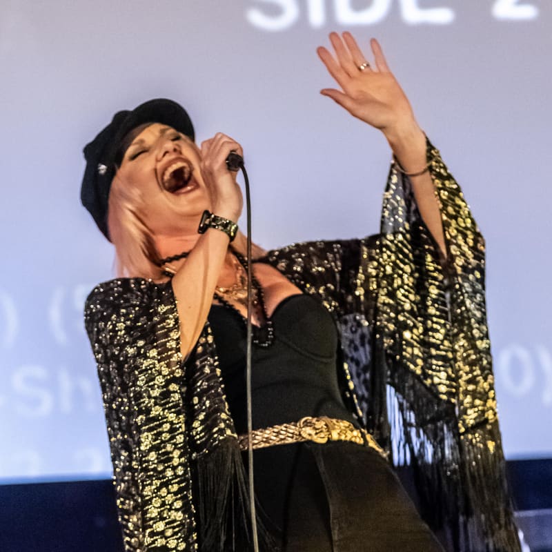 Performer in a sequinned gold and black jacket singing expressively with arm raised on stage.