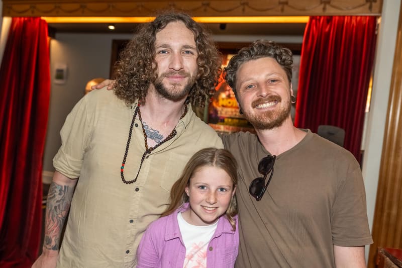 Three people smile at the camera in front of red velvet curtains at an indoor venue.