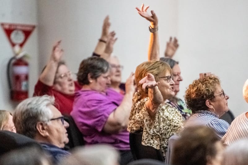 Audience members raise their hands and arms during an interactive moment at a gallery event, with white walls and artwork visible in the background.