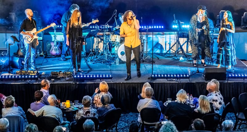 Band performing on stage with blue lighting, featuring a vocalist in yellow and musicians with guitars and keyboards.