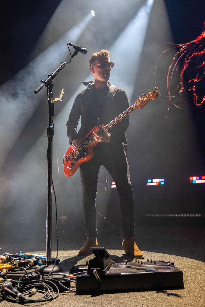 Guitarist performing on stage with a red bass guitar under dramatic white spotlights during a Royal Blood concert.
