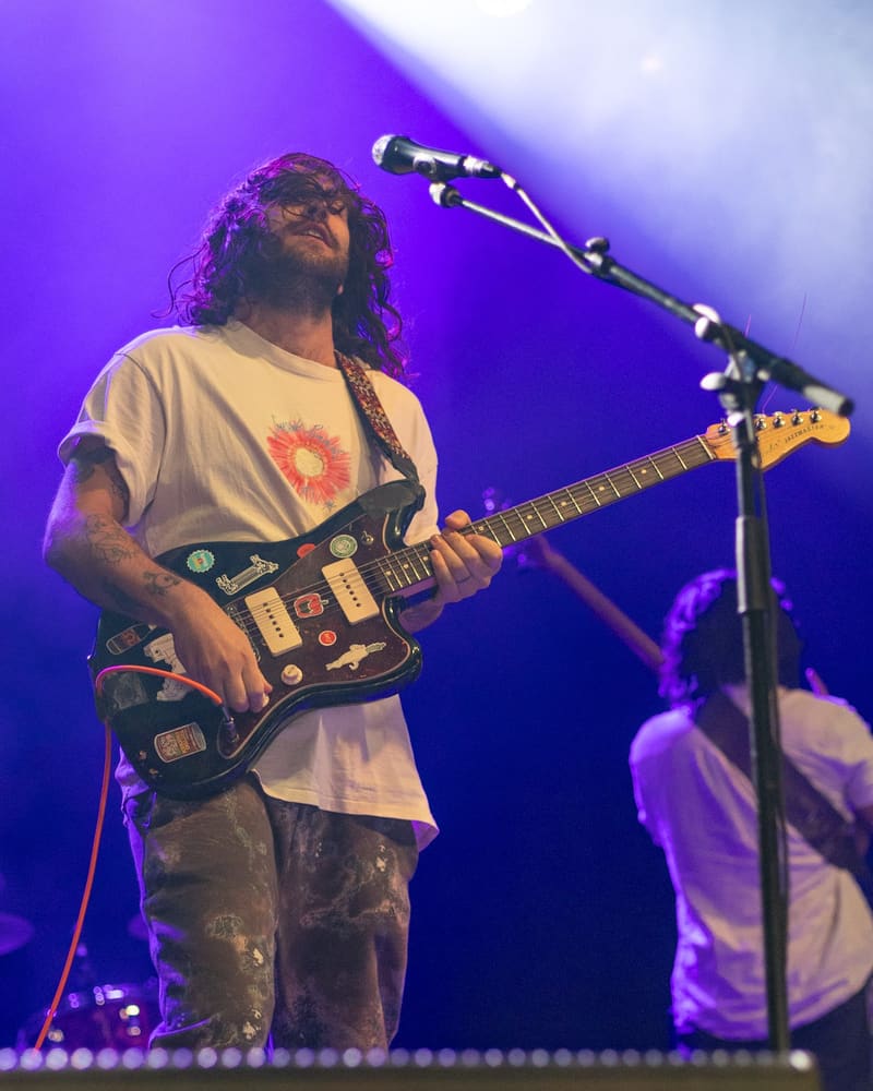 Guitarist with long dreadlocks performing on stage under purple lighting during a rock concert.