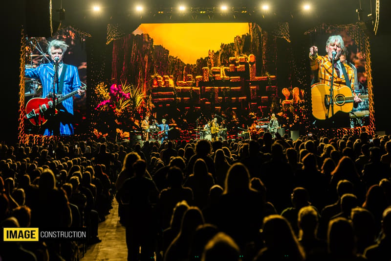 Two musicians perform on stage before a large crowd illuminated by warm yellow and orange lighting during a concert.
