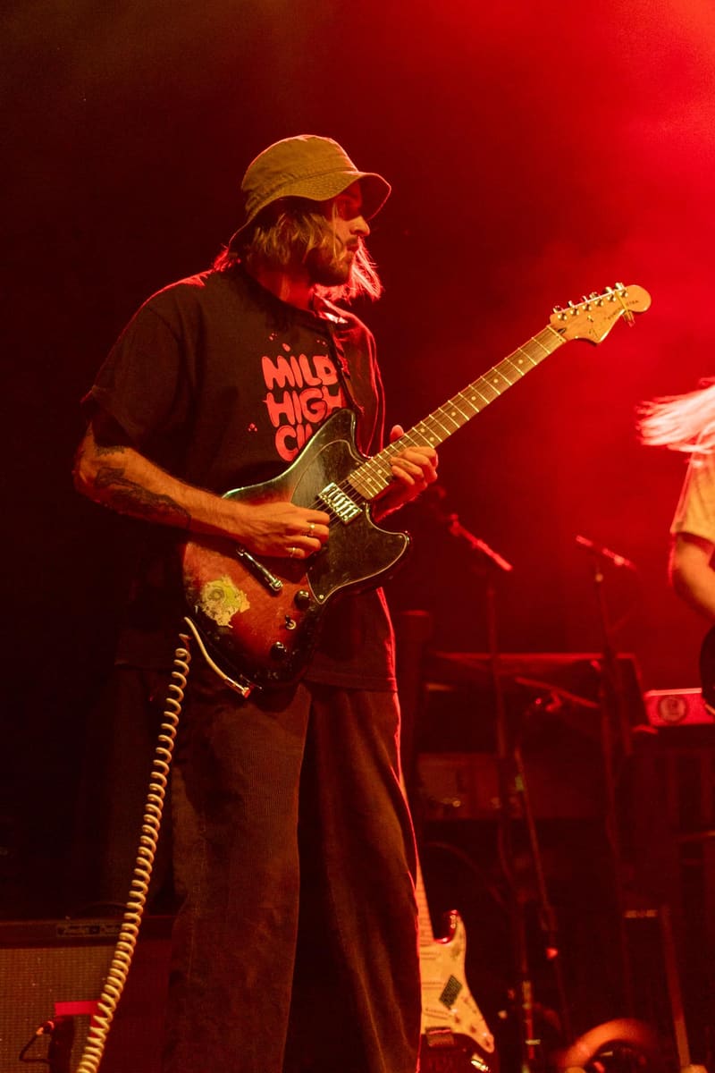 Guitarist performing on stage bathed in red light, wearing a cap and graphic t-shirt while playing an electric guitar.
