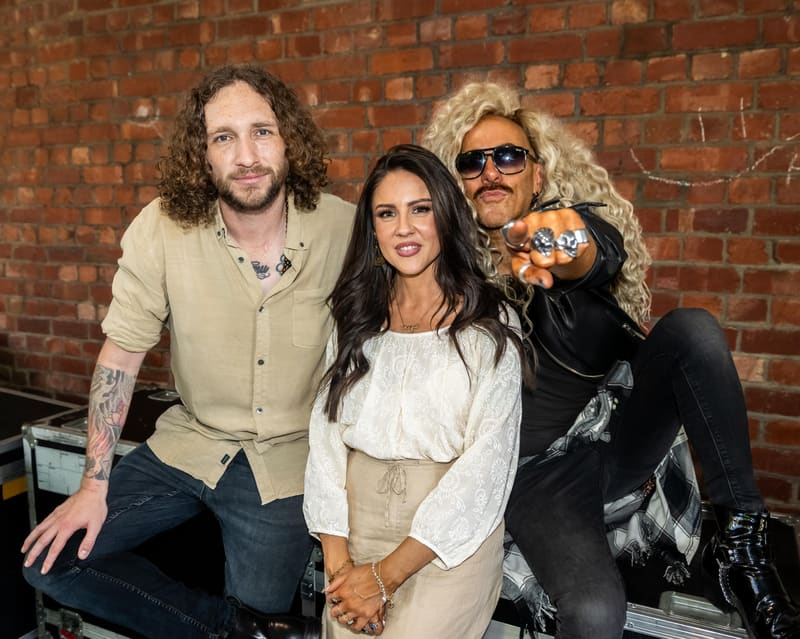 Three people pose together against an exposed brick wall in a gallery setting.