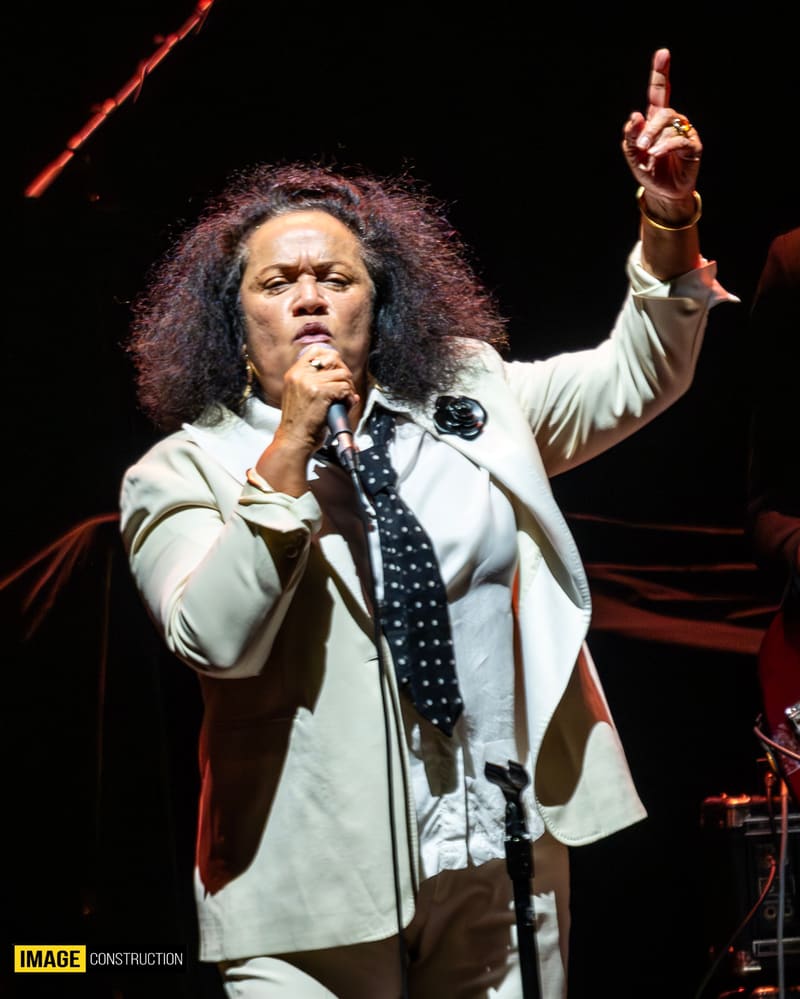 Performer at microphone in cream blazer and tie, pointing upward with one hand during stage performance.