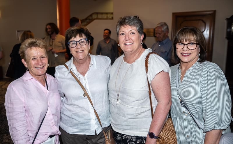 Four women pose together at a gallery opening, smiling at the camera in an indoor social setting.