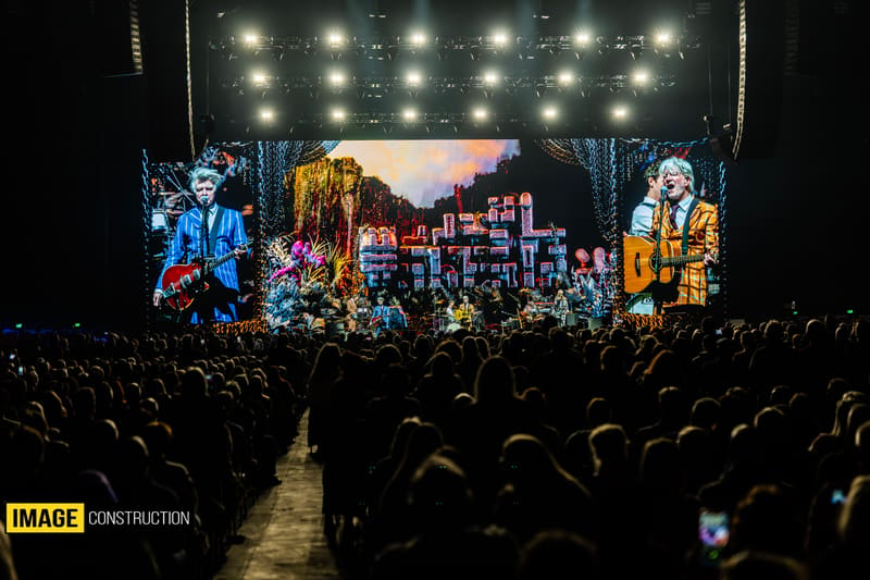 Large concert stage with illuminated band members and cityscape projection, crowd silhouetted in foreground.