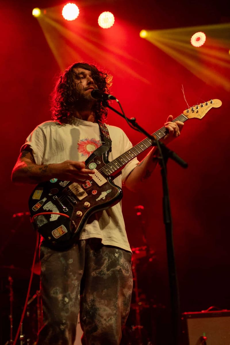 Guitarist performing at microphone under red stage lighting with electric guitar.