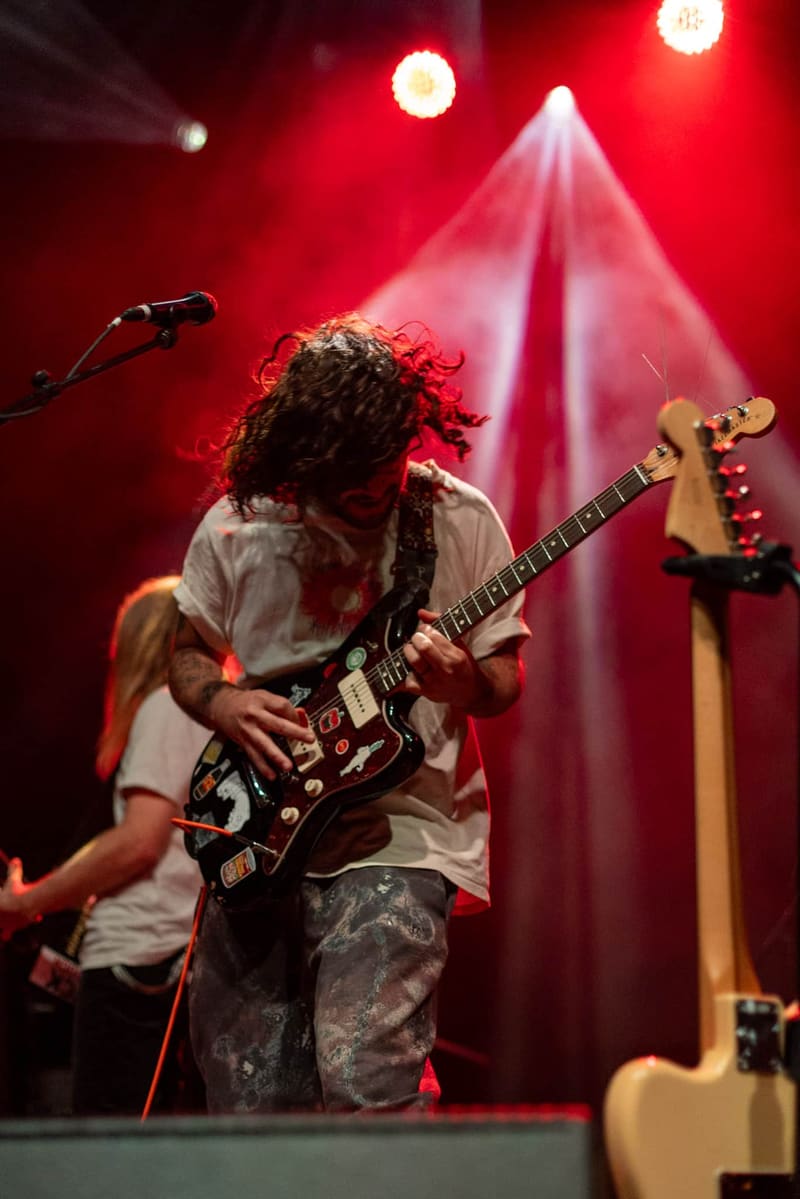 Guitarist with long dark hair performing on stage bathed in red and pink stage lighting.