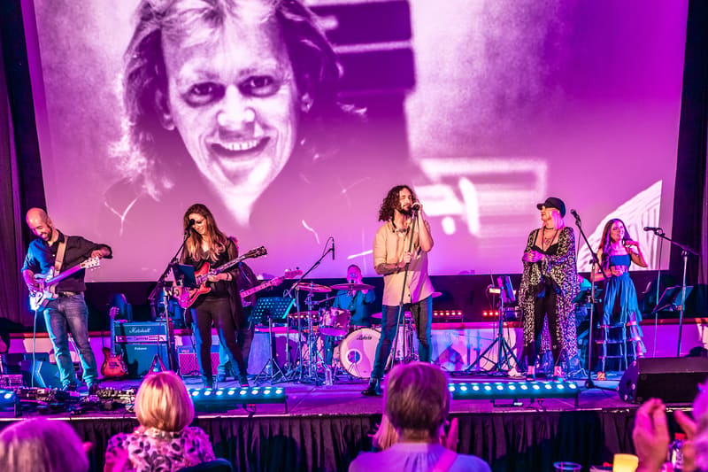 Live band performs on stage beneath a large portrait screen, bathed in purple and blue stage lighting.