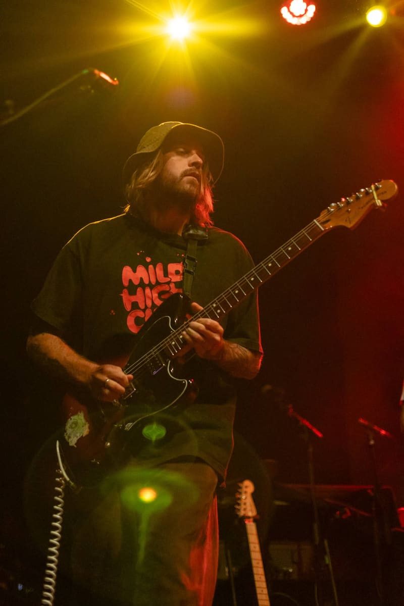 Guitarist in a black hat and red-text shirt performing under warm stage lighting with electric guitar.