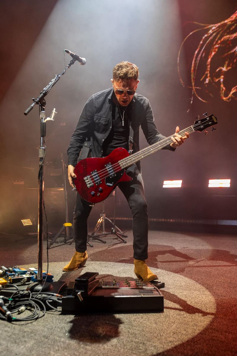 Bassist in dark jacket and red shoes plays electric bass guitar under stage spotlights during a live performance.