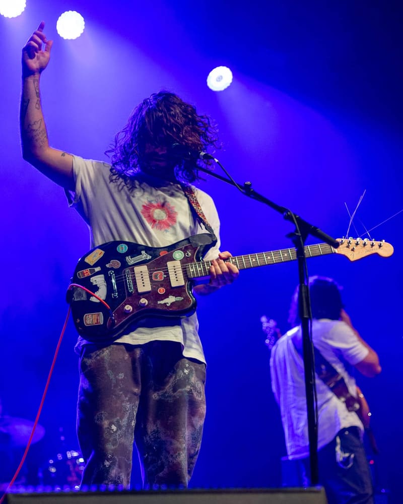 Guitarist with long dark hair performs at a microphone under purple stage lights, raising one fist while playing an electric guitar.