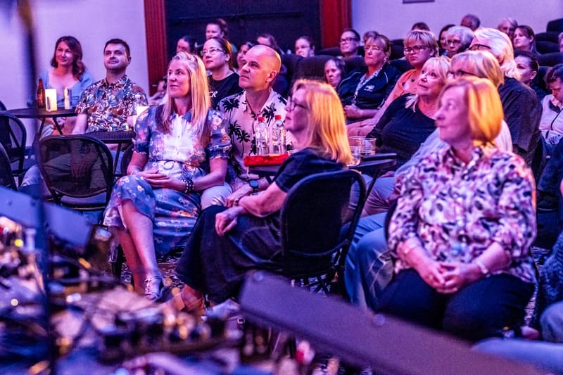 Seated gallery visitors watch a performance in warm purple and pink stage lighting.