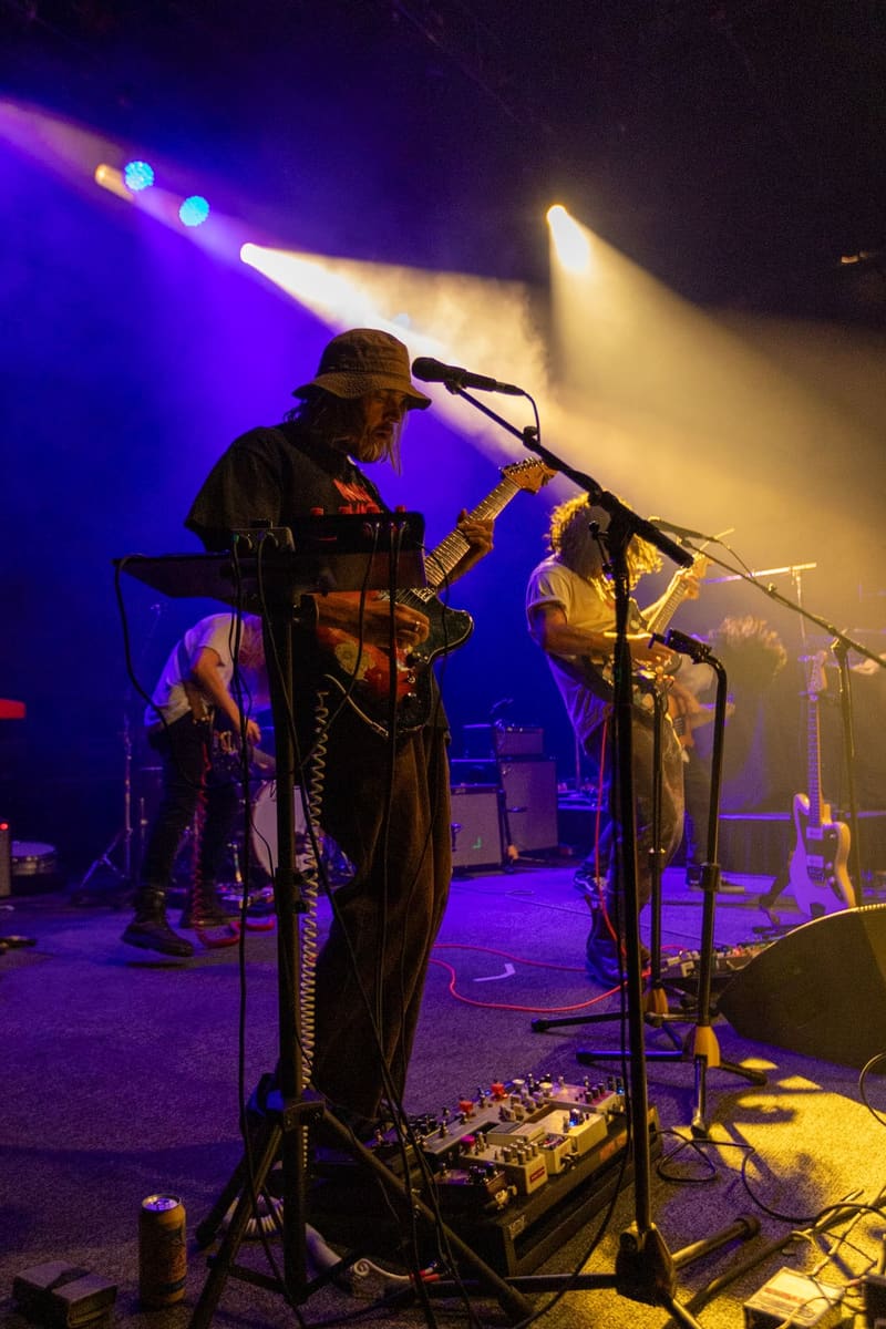 Saxophonist in hat performing at microphone under purple and yellow stage lights with band members visible behind.