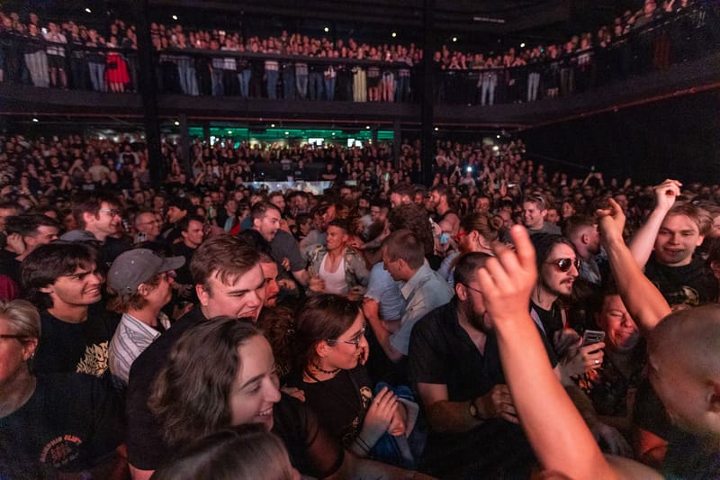 Crowded concert venue with audience members raising arms and cheering during a live rock performance.