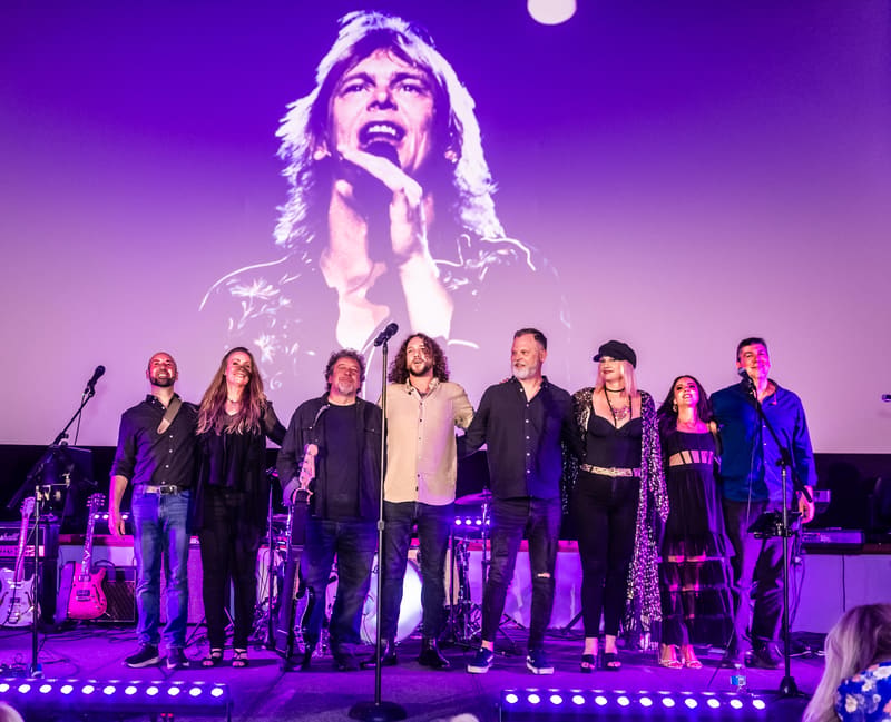 Group of performers standing on stage beneath a large projected image, lit by purple stage lighting.