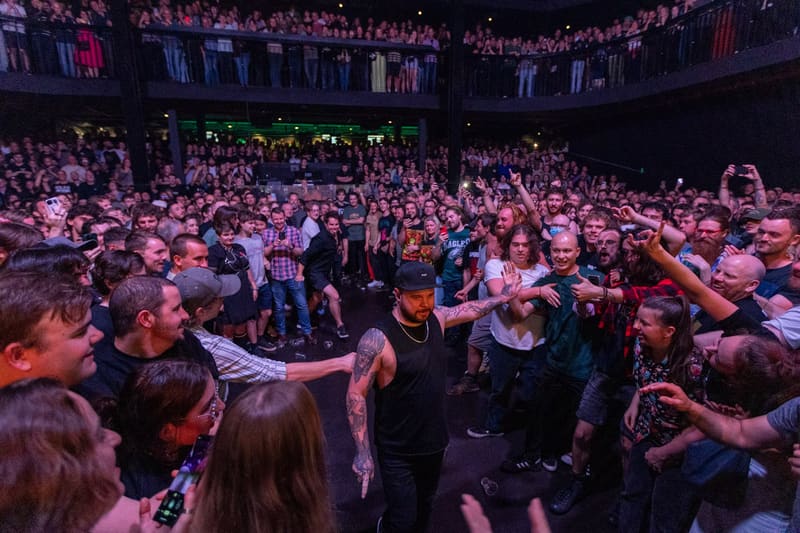 Large crowd of enthusiastic fans with raised arms surrounding performers on stage at a concert venue.