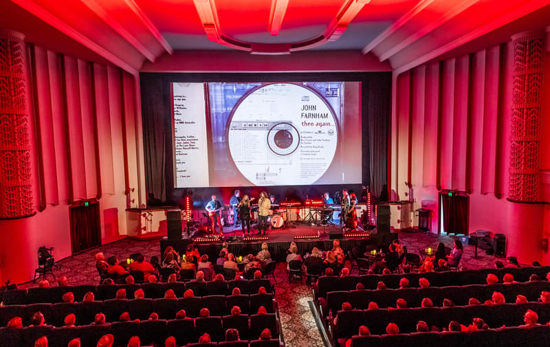 Large theatre audience seated facing a stage lit in red, with performers visible and a CD image projected on the screen above.