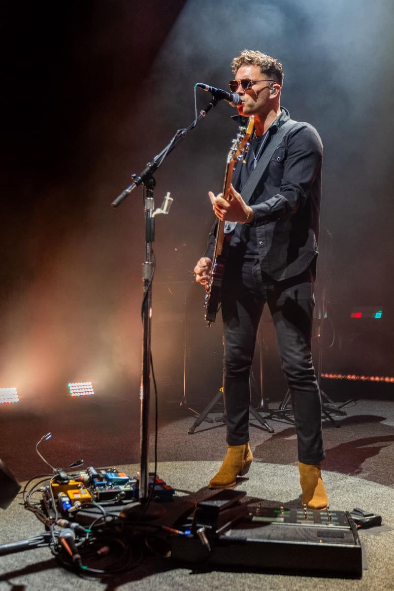 Guitarist in dark suit and yellow boots performing at microphone under warm stage lighting.