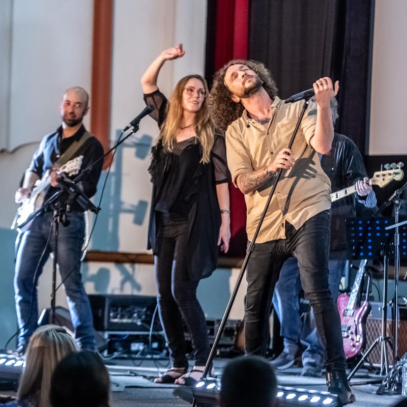 Four musicians performing on stage, with a vocalist centre-stage and guitarists flanking left and right, audience members visible in foreground.