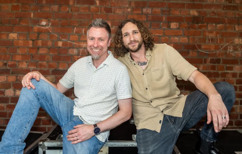 Two men pose together against an exposed brick gallery wall, one in a light blue shirt and jeans, the other in a tan cardigan.