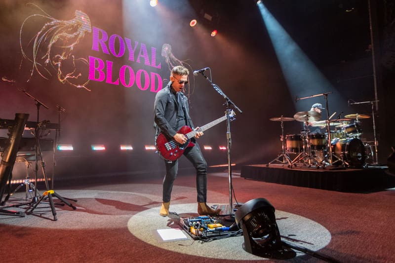 Guitarist performing on stage under pink and blue stage lights with "Royal Blood" text projected behind.