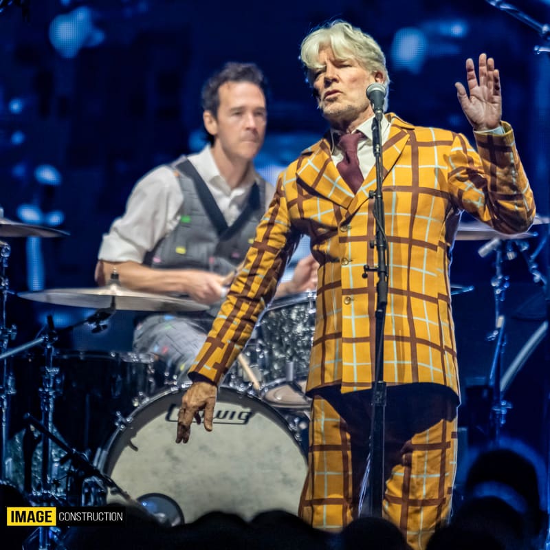 Two musicians perform on stage, one in a bold yellow tartan jacket gesturing expressively while another plays drums in the background.