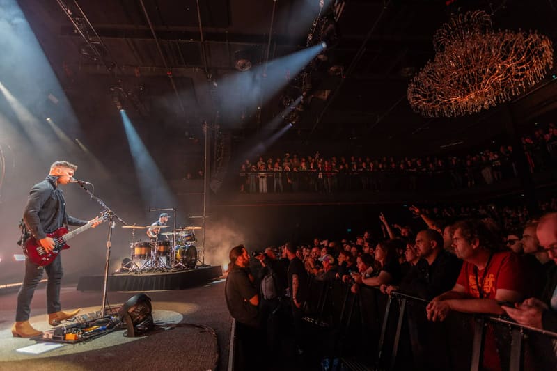 Guitarist performs on stage with drummer and crowd bathed in red and blue stage lighting.