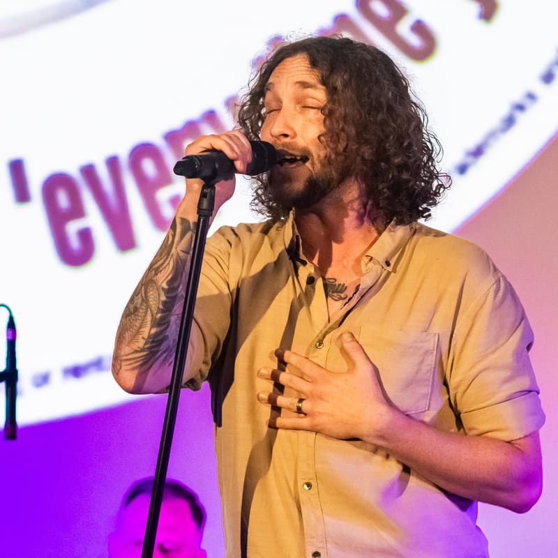 Performer with long curly hair singing at a microphone, wearing a yellow shirt under purple stage lighting.