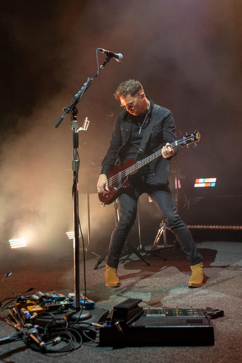 Bassist in dark jacket performs on stage under warm spotlights, focused on instrument with microphone stand nearby.