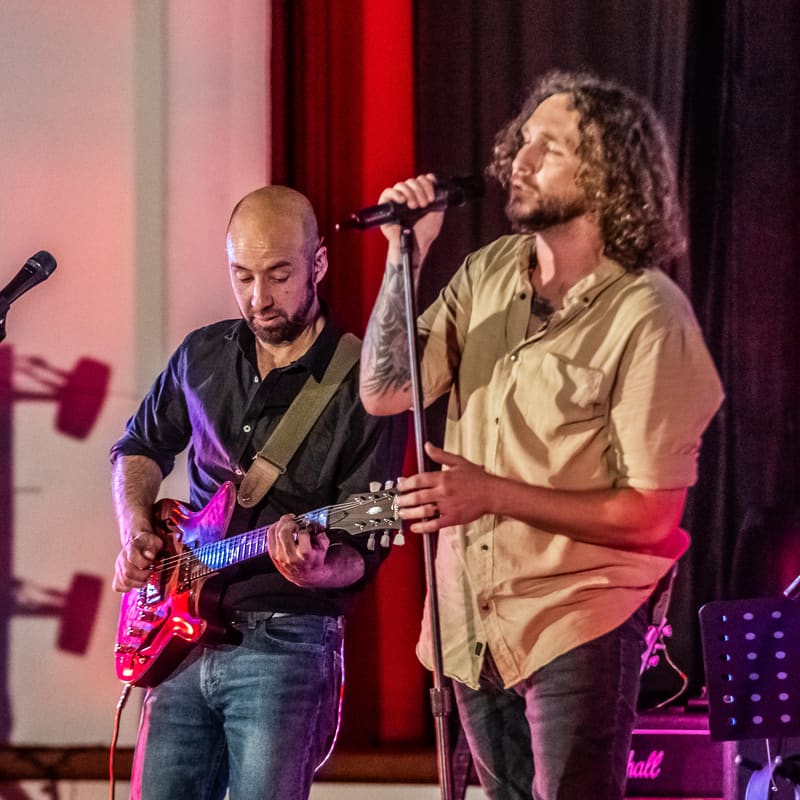 Two musicians perform on stage, one playing electric guitar and the other singing into a microphone, lit by red and purple stage lighting.